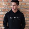 A young man with short dark hair smiles and looks to the side while standing against a brick wall, wearing a black hoodie with "FRIENDS" printed in the show's iconic font on the front.