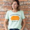 A woman stands against a brick wall, smiling and looking to the side. She wears a light green T-shirt with an orange couch graphic and the text "I'D RATHER BE WATCHING FRIENDS.