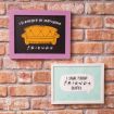 Two framed signs hang on a brick wall. The top one has a yellow couch and says "I'd rather be watching Friends." The bottom one has a speech bubble and says "I speak fluent Friends quotes.