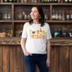 A woman stands in front of wooden shelves filled with glassware, wearing a white t-shirt with images of beer glasses and bottles and the text "I DO CRAFTS." She is smiling and looking to the side.