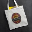 A white tote bag with a colorful round "Butterbeer" logo on the front. The bag contains a yellow folder and a brown notebook, and is placed on a dark surface.