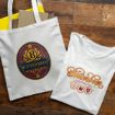A white tote bag with a colorful "Butterbeer" logo and a white t-shirt with a decorative "Butterbeer" design are displayed on a wooden surface. The tote bag handles contain yellow items.
