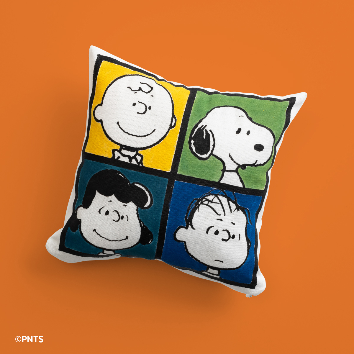 A square pillow featuring illustrated portraits of Charlie Brown, Snoopy, Lucy, and Linus from the Peanuts comic, each in a colored square, on an orange background.