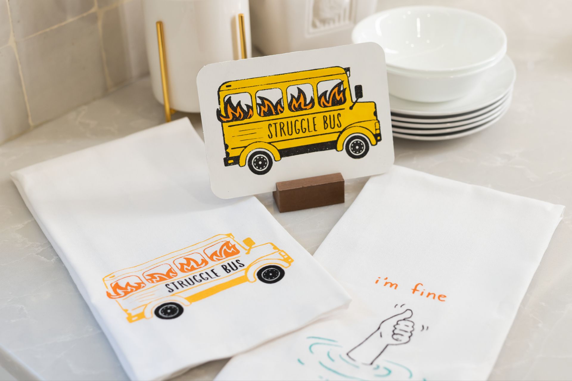 A countertop displays two white towels, one with a yellow bus labeled "Struggle Bus" and another with a hand giving a thumbs-up in water with the words "i'm fine." A matching "Struggle Bus" card stands nearby.