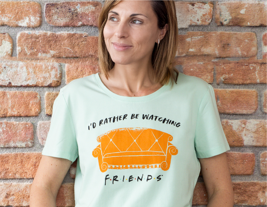 A woman with light brown hair stands against a brick wall, smiling slightly. She is wearing a light green T-shirt with an orange couch graphic and the text "I'd rather be watching FRIENDS.