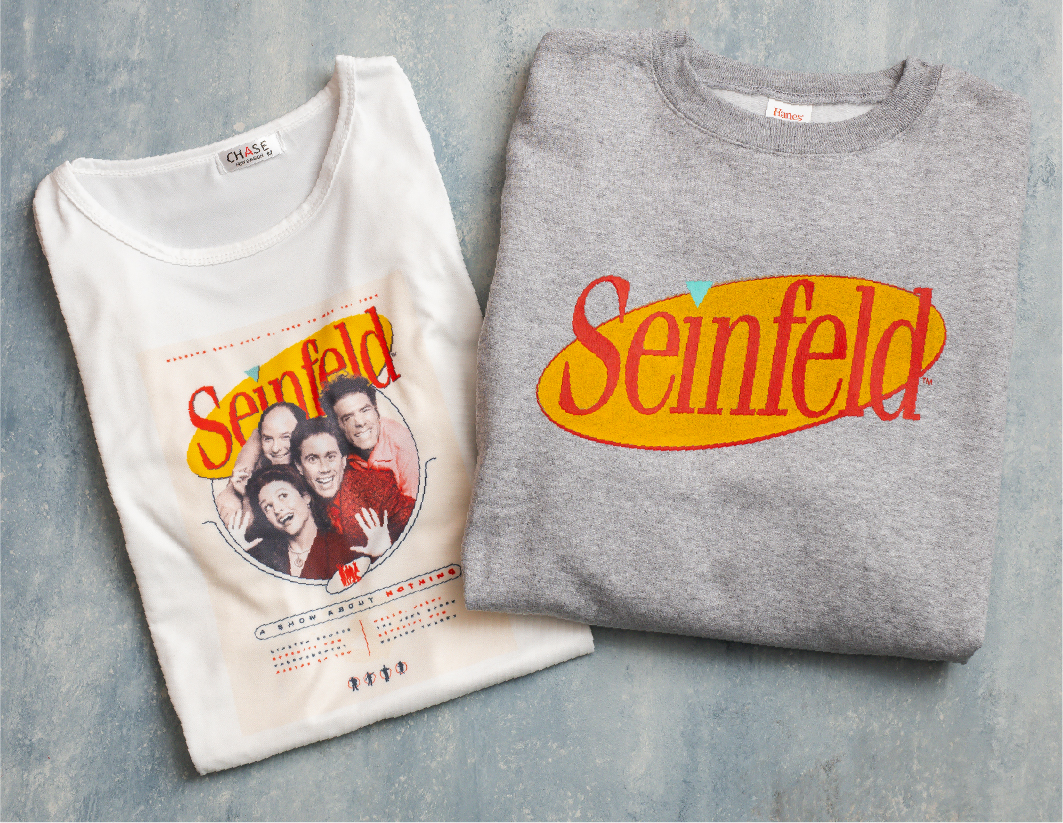 Two folded shirts on a light surface: one white with a Seinfeld cast photo and logo, and one gray with a large red and yellow Seinfeld logo.
