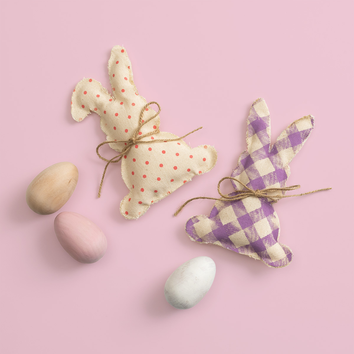 Two fabric bunny decorations, one with red polka dots and the other with purple checkered pattern, both tied with twine bows, are placed on a pink background next to four pastel-colored eggs.