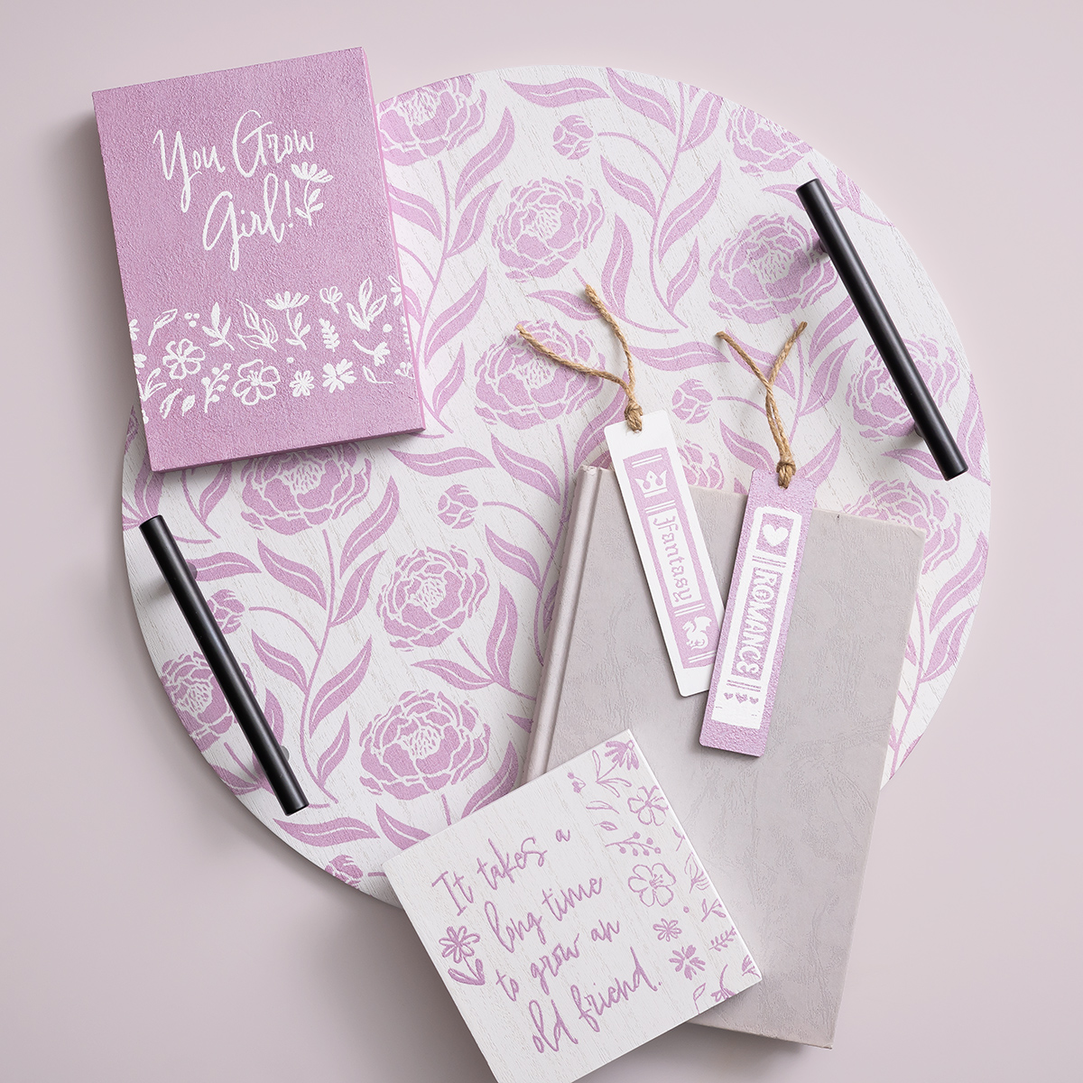A round tray with a purple floral pattern holds a purple notebook labeled "You Grow Girl!", two bookmarks with positive messages, a beige notebook, and a white card with a friendship quote, all on a light pink background.