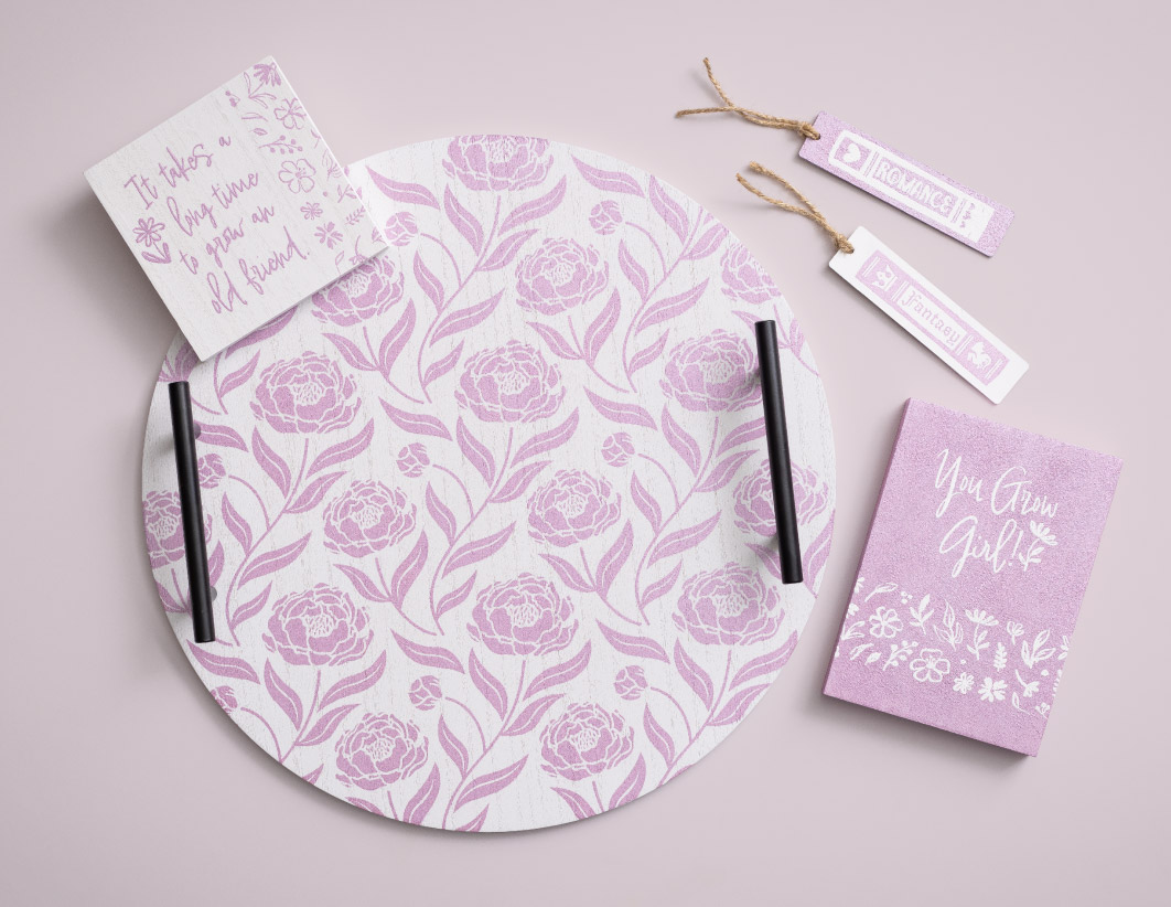 A set of pink and white floral-themed stationery on a pastel pink background, including a round tray, a notebook, a notepad, and two bookmarks with inspirational quotes and floral patterns.