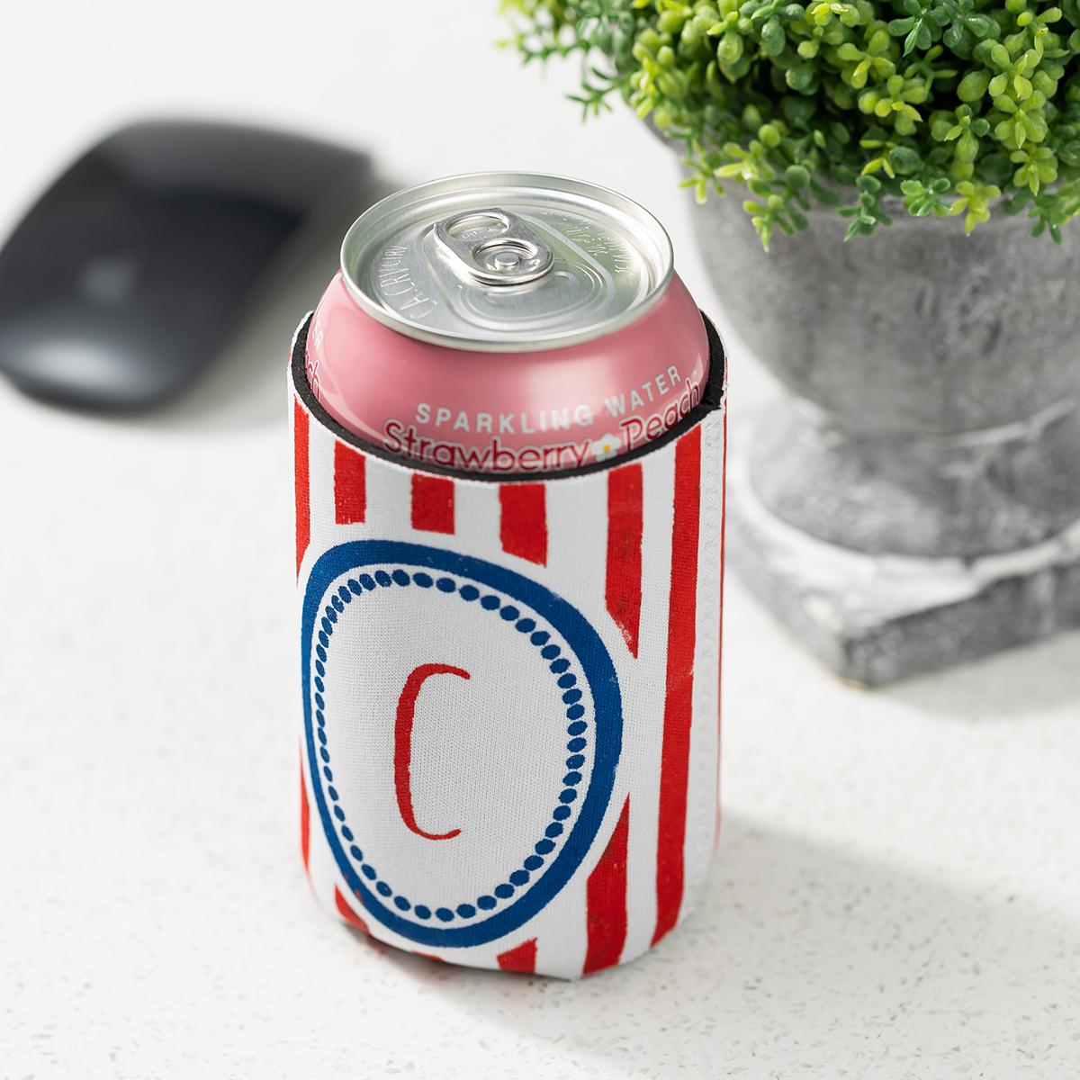 A can of sparkling water in a red and white striped koozie with a blue-bordered circle and the letter "C," next to a gray computer mouse and a potted green plant on a white desk.