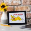 A framed picture of Disney’s Pluto lying down and smiling, with the text "I LOVE MY DOG (AND LIKE THREE PEOPLE)" above him. The frame is on a desk next to a vase with a sunflower and a yellow pencil.