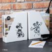 Two white magazine holders with black sketch illustrations of Minnie Mouse and Mickey Mouse sit on a desk against a brick wall, alongside office supplies like a notepad, pencil, stapler, and a lamp.
