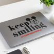 A laptop with a sticker showing Mickey Mouse’s face and the words "keep smiling (it confuses people)" in black and red text. A potted plant and a cup are nearby on the desk.