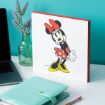 A decorative block featuring Minnie Mouse in a red dress and bow stands on a desk beside a turquoise notebook, pencil, glass vase with lavender, and a laptop, against a bright teal wall.