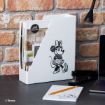 A white magazine holder with a black sketch of Minnie Mouse stands on a black desk beside a stapler, notepad, pen, and a laptop, against a brick wall background.