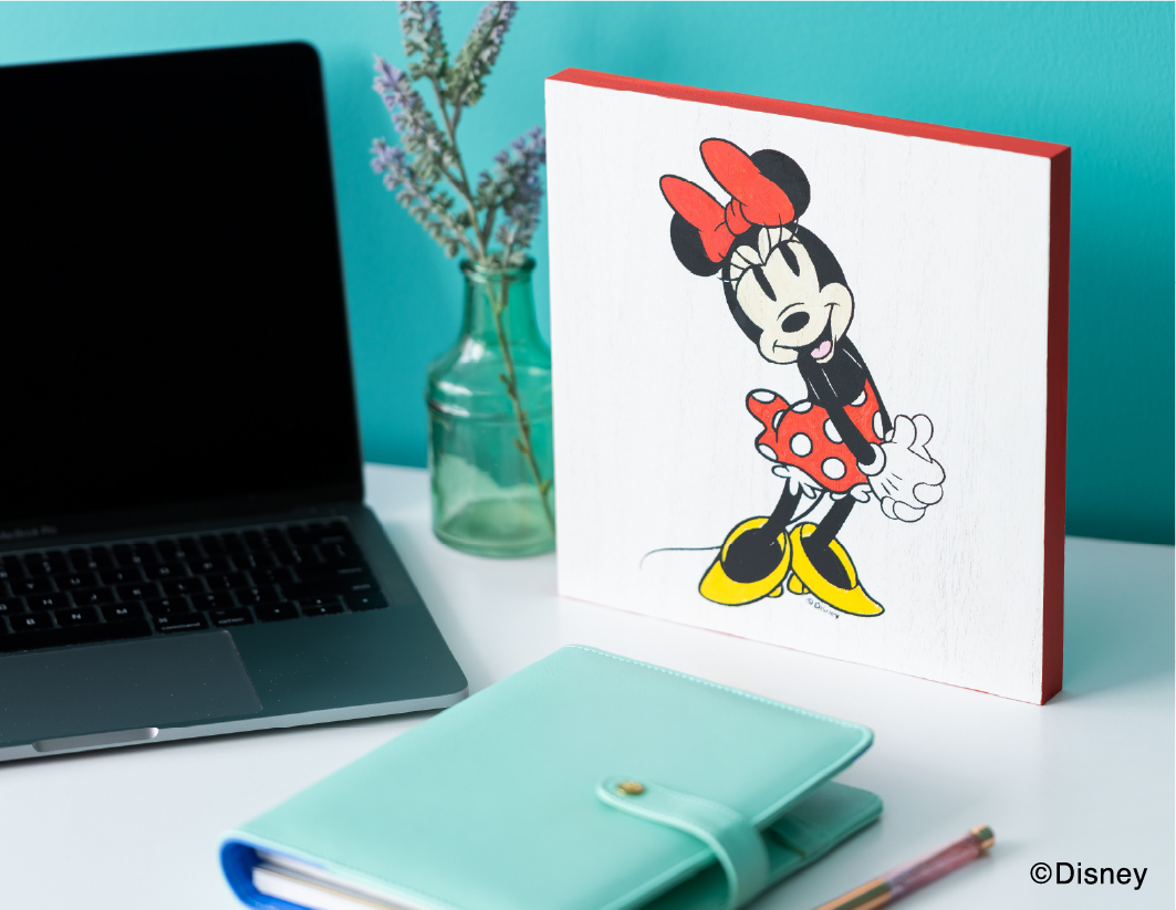 A desk with a laptop, an aqua notebook, a small glass vase with lavender, and a Disney artwork of Minnie Mouse in a red polka-dot dress and yellow shoes against a teal wall.