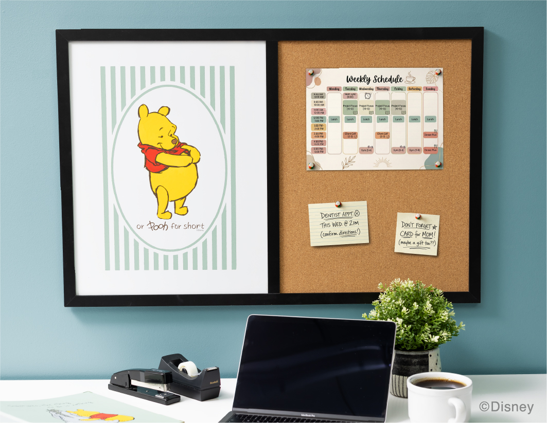 A desk with a laptop, coffee, and plant sits below a framed board featuring a Winnie the Pooh illustration on the left and a weekly schedule with sticky notes pinned on corkboard to the right.