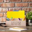 A beige desk organizer with Winnie the Pooh character faces holds colorful file folders, pens, pencils, and markers. A stapler and a small potted plant sit nearby on a wooden desk against a brick wall.
