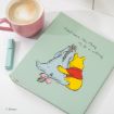 A light green notebook with Winnie the Pooh hugging Eeyore and the text "Sometimes, the thing to do is nothing" on the cover. Next to it is a pink cup of coffee and a light blue pen on a white surface.