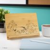 A wooden tablet stand engraved with line drawings of Tigger, Eeyore, Piglet, and Winnie the Pooh. The stand is on a desk with a plant, a mug, and a closed tablet nearby.