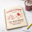 A spiral notebook with a cover illustration of Winnie the Pooh pulling a wagon of honey pots, labeled "Pooh's Farm," beside a pink mug of hot chocolate with whipped cream and an open notepad on a white desk.