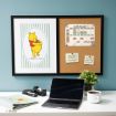 A workspace with a laptop, coffee mug, stapler, plant, and notebook sits below a wall frame. The frame displays a Winnie the Pooh illustration and a corkboard with a weekly schedule and notes pinned to it.