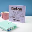 A lavender book titled "Relax: Take a moment Eeyore" with an illustration of Eeyore lying on his back, sits on a white surface next to a pink mug of coffee and a mint-green planner.