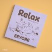 A square sign with an illustration of Eeyore lying on his back surrounded by flowers. The text reads, “Relax, Take a moment, EEYORE.” The background is light purple, and the sign is set on a yellow surface.