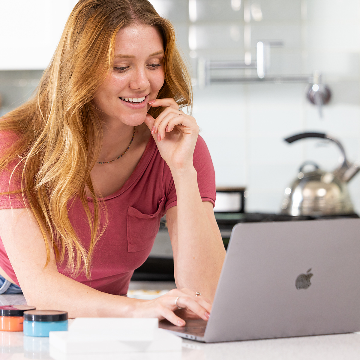 A woman with long red hair, wearing a pink t-shirt, smiles while using a laptop in a bright kitchen. She is leaning on a counter with paint containers and paper nearby.