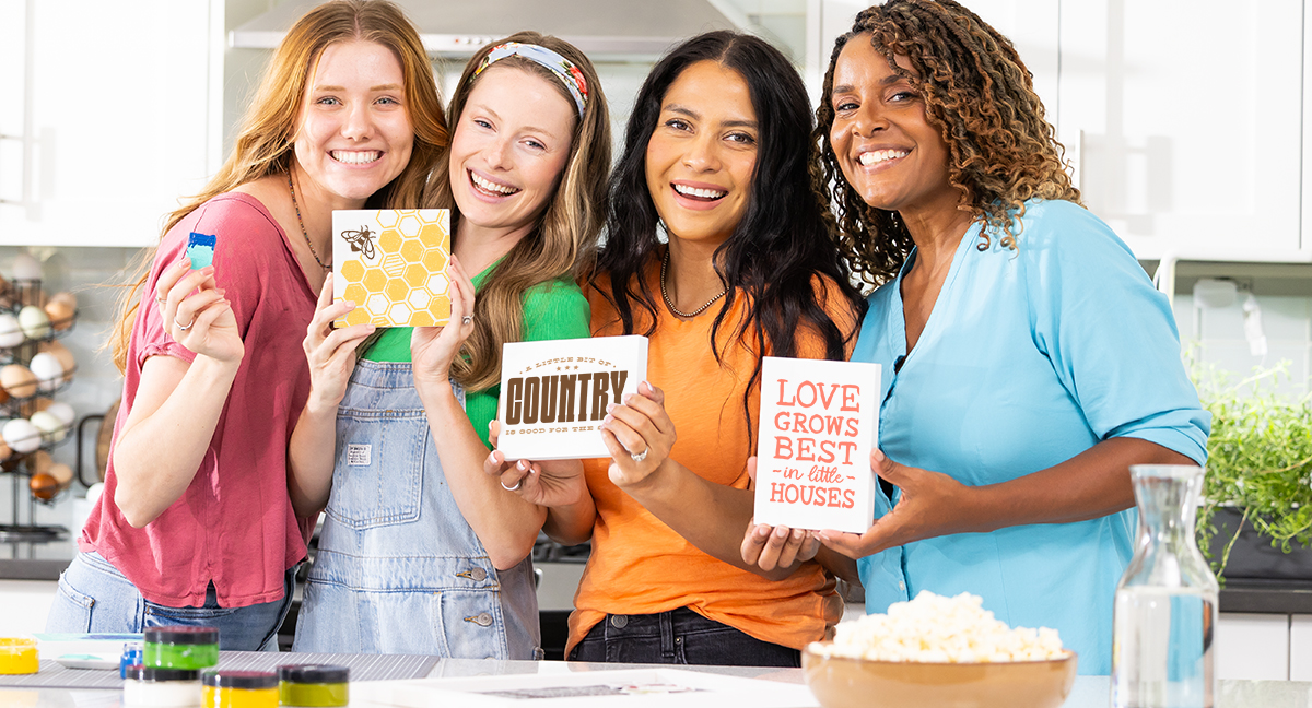 Four smiling women stand together in a bright kitchen, each holding a colorful decorative sign with positive or inspirational messages. A bowl of popcorn and craft supplies are on the counter in front of them.