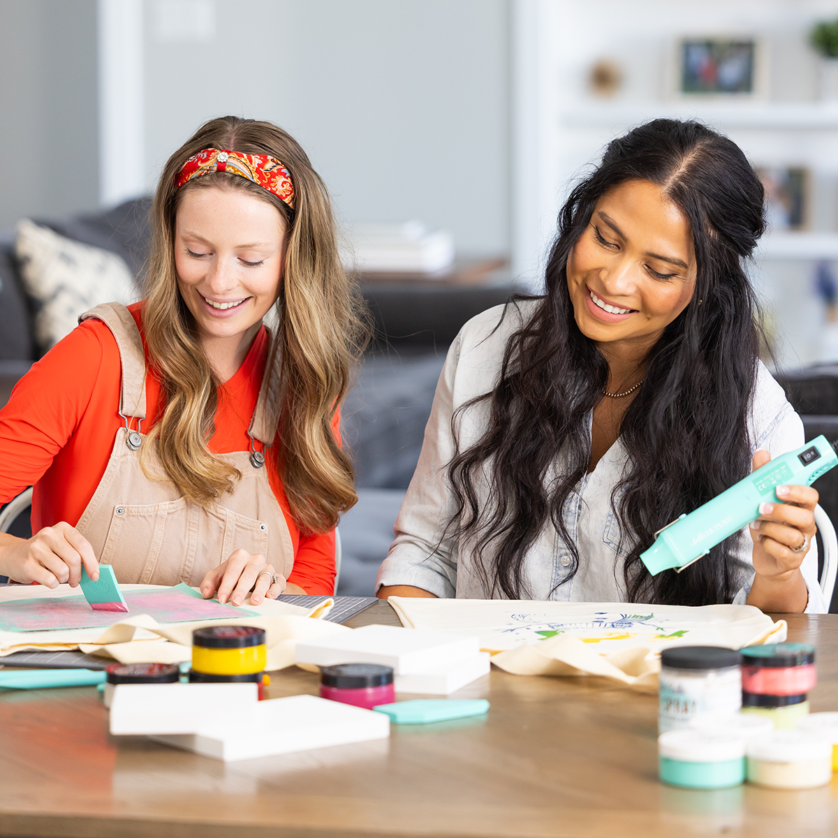 Two women sit at a table smiling while working on a craft project with colorful materials, including jars of paint, brushes, and paper. The background shows a cozy, modern living room.