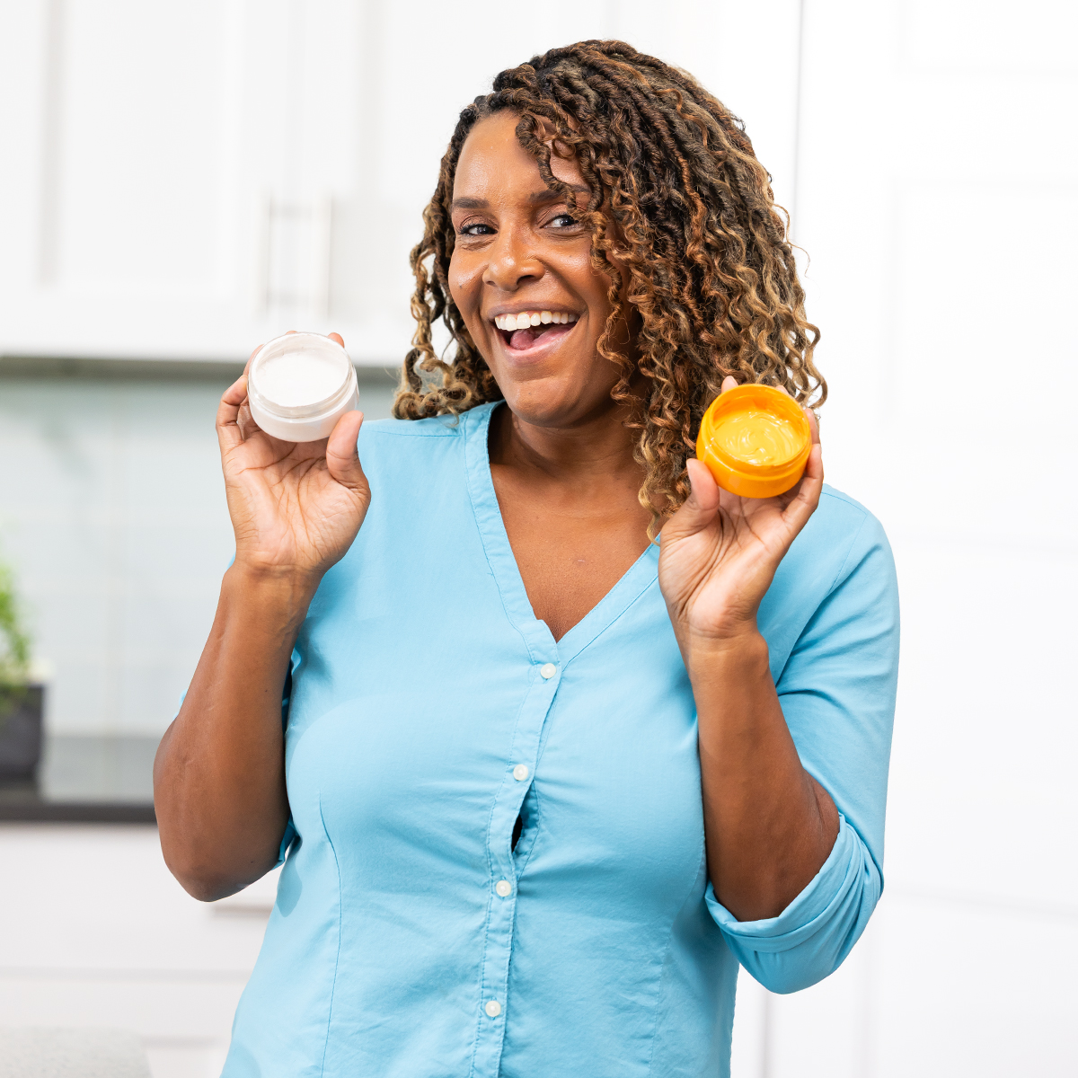 A smiling woman in a light blue shirt holds two open jars of cream, one white and one yellow, in a bright kitchen.