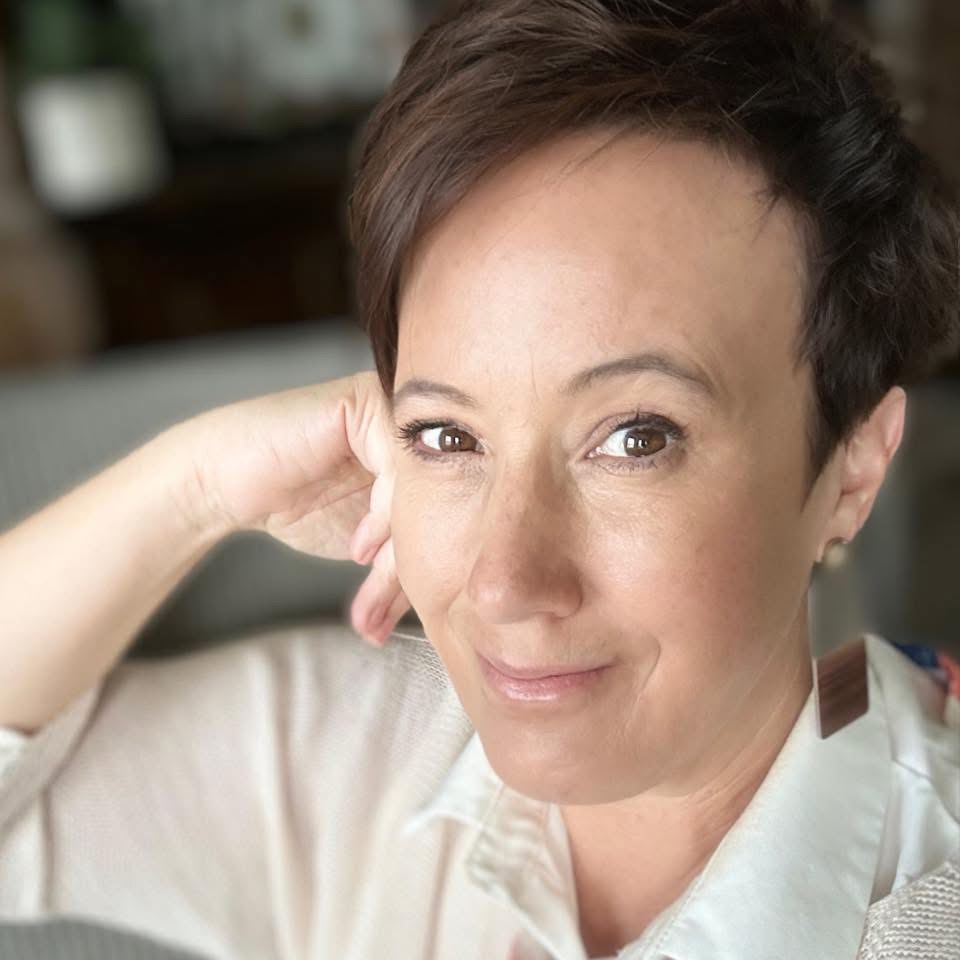 A person with short brown hair and light skin smiles softly at the camera, resting their chin on their hand. They are wearing a light-colored collared shirt and large earring, with a blurred background behind them.