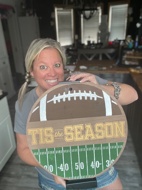 A woman smiling and holding a round football-themed sign that says "TIS the SEASON" with a football field design at the bottom, standing in a kitchen with light from windows behind her.