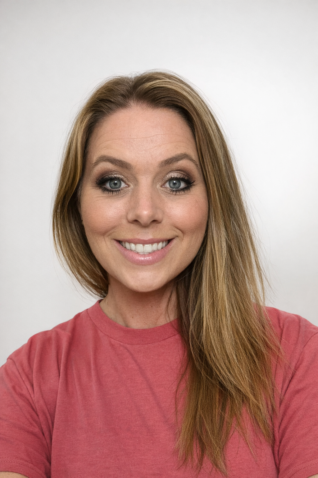 A woman with long, straight, light brown hair and blue eyes smiles at the camera. She is wearing a pink t-shirt and has natural makeup. The background is plain and light-colored.
