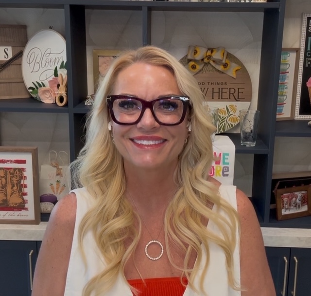 A smiling woman with long blonde hair and dark-rimmed glasses sits in front of a shelf decorated with framed art, flowers, and decorative signs. She is wearing a white top over a red shirt and a circular necklace.