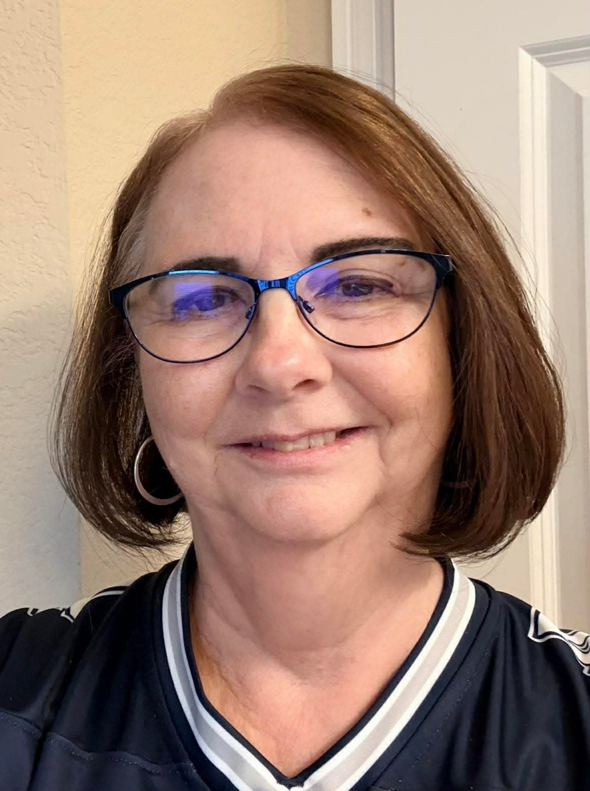 A woman with short brown hair and glasses smiles at the camera. She is wearing a dark top with a white collar and a hoop earring, standing indoors against a light-colored wall and door.