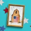 A framed painting of a brown barn with an American flag on its front, set against a blue background with red, white, and blue star-shaped decorations around it.