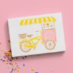 A card with a drawing of a yellow ice cream bike cart, featuring a striped canopy and the words "Sunny with a chance of sprinkles," is set on a pink background with colorful sprinkles scattered nearby.