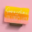 A sign with yellow, orange, and pink paint strokes on a pink background reads, "Summertime and the livin’s easy," in white cursive and bold letters, supported by a small wooden stand.