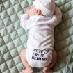 A baby in a white hat and onesie lies on their stomach on a quilted mint-green blanket, with the words "Cute From Behind" and a heart symbol printed on the back of the onesie.