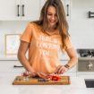 A woman in a peach t-shirt that says "JESUS LOVES YOU & I'm Trying" smiles while slicing strawberries on a wooden cutting board in a bright, modern kitchen. Blueberries and a small dish are on the counter.
