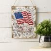 A rustic, vintage-style sign featuring a waving American flag hangs on a white wooden wall above a shelf with a black-and-white polka dot planter holding a green leafy plant. Handwritten script decorates the sign.