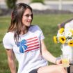 A woman sits on grass holding a cup of lemon water. She wears a white "All American Girl" t-shirt with a blue bow and red stripes, and there is a bouquet of sunflowers in the background.