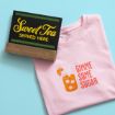 A pink t-shirt with an illustration of iced tea and the words "Gimme Some Sugar" next to a black sign with "Sweet Tea Served Here" in yellow and green letters on a light blue surface.