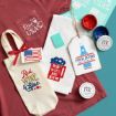 A flat lay of patriotic-themed gifts and craft supplies, including a red shirt, printed towel, bottle tote, painted ceramic tag, American flag patch, ink jars, and stencils, all in red, white, and blue colors.