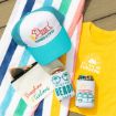 A striped beach towel with sunglasses, a "Sunshine & Tanlines" pouch, a turquoise "Tanned & Tipsy" hat, a yellow "You Are My Sunshine" shirt, and two can coolers with beach designs.