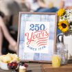 A framed sign on a table reads "250 Years Since 1776" in red and blue, with stars and stripes. The table holds flowers, lemonade, snacks, and utensils. People are gathered and blurred in the background.