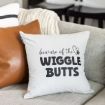 A white throw pillow on a gray couch with the words "beware of the WIGGLE BUTTS" and a small paw print graphic. A brown pillow and a cream knit blanket are also visible.