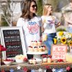 A woman wearing a "250 Years Since 1776" shirt stands by a table with patriotic cupcakes, fruit, sunflowers, a framed patriotic poem, and a "1776 America" sign at an outdoor celebration with others in the background.